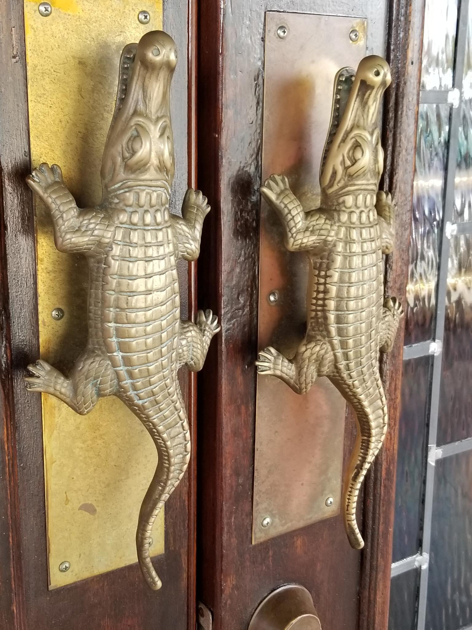 A pair of door handles made of brass in the shape of either alligators or crocodiles on wooden doors in which part of a tiled-glass panel is visible.