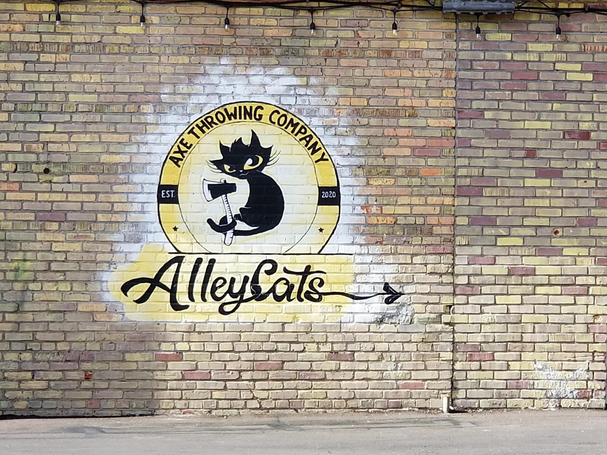 A mural on a brick wall advertising the Alley Cats Axe Throwing Company and featuring a cartoon black with large eyes kitten holding an axe.