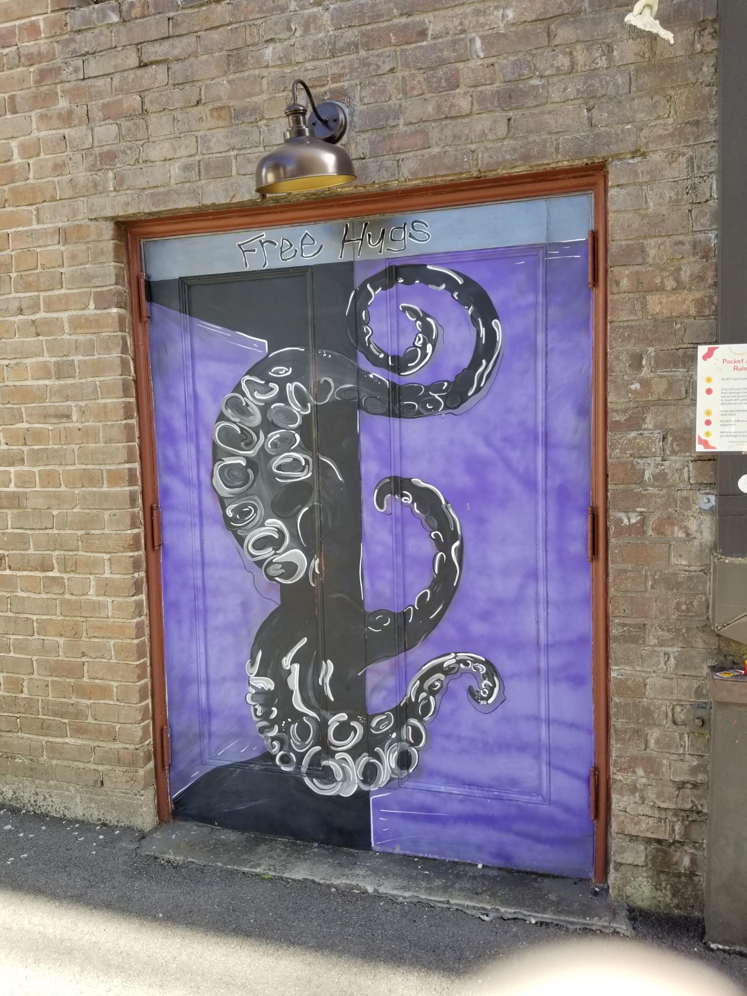 A pair of exterior doors painted purple, bearing the words 'Free Hugs' and made to appears as if one door is open and a giant octopus is reaching out from beyond.