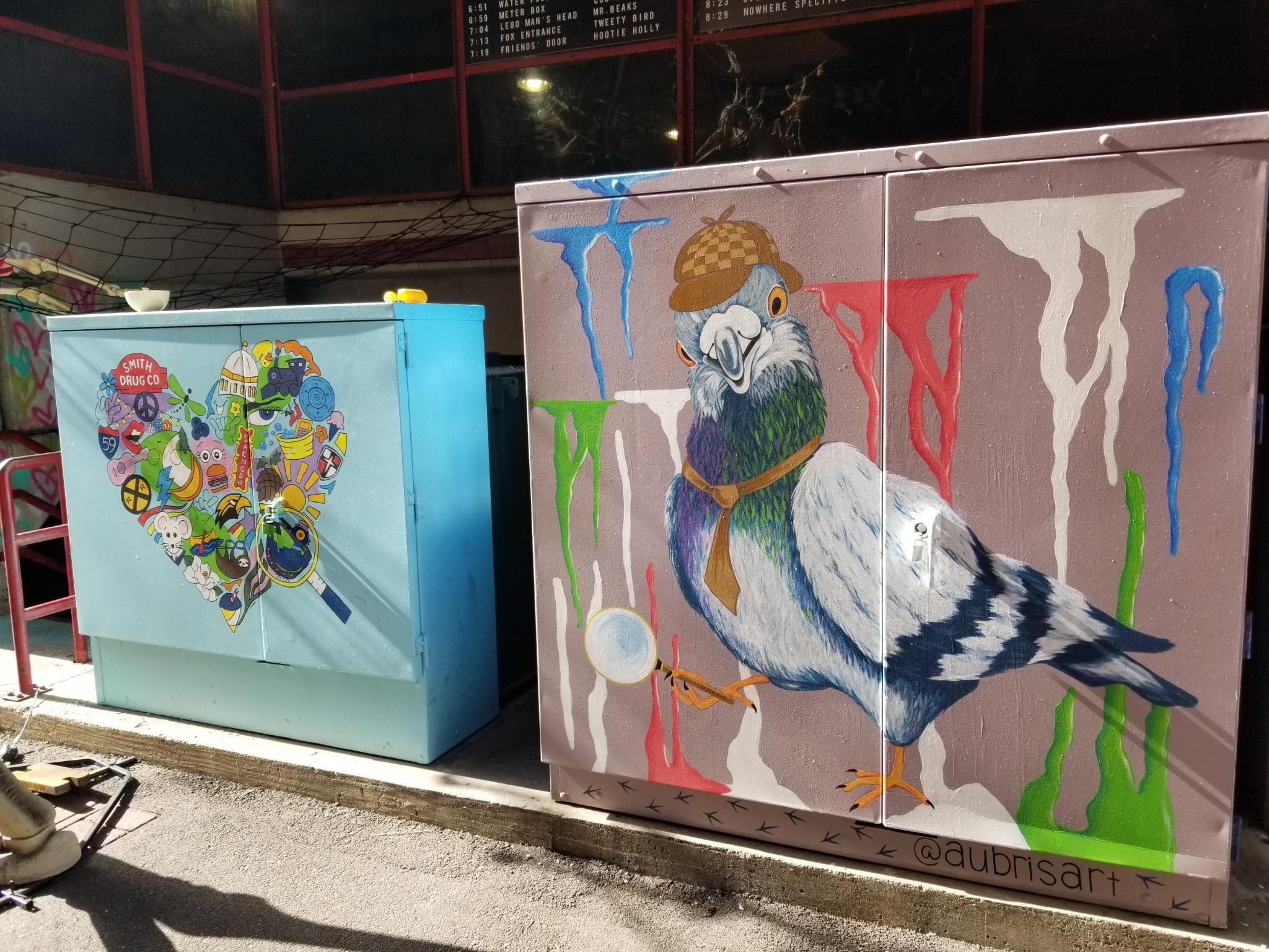 Two utility boxes carrying paintings. The closer one portrays a large pigeon wearing a Sherlock Holmes style hat and tie, carrying a magnifying glass, and multicolored splotches in the background seem to signify bird droppings. It is signed @aubrisart. The smaller further box portrays many elements inside a heart shape.