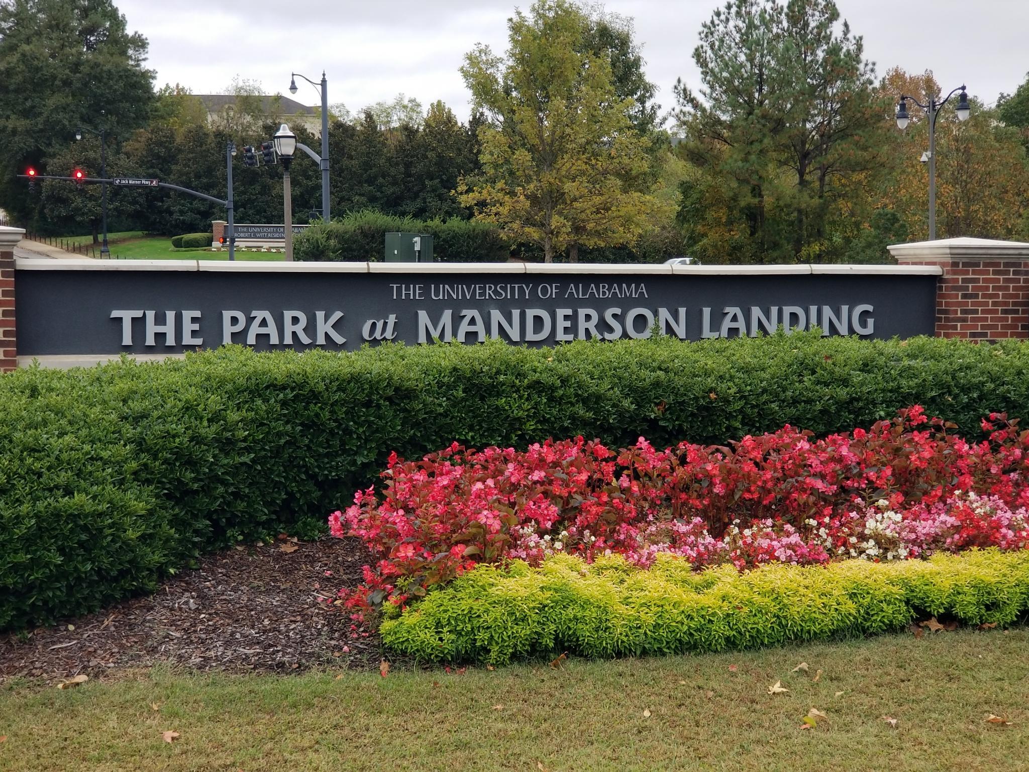 A sign indicating the location of 'The University of Alabama, The Park at Manderson Landing', with a flower bed visible in front of it and the university visible behind.