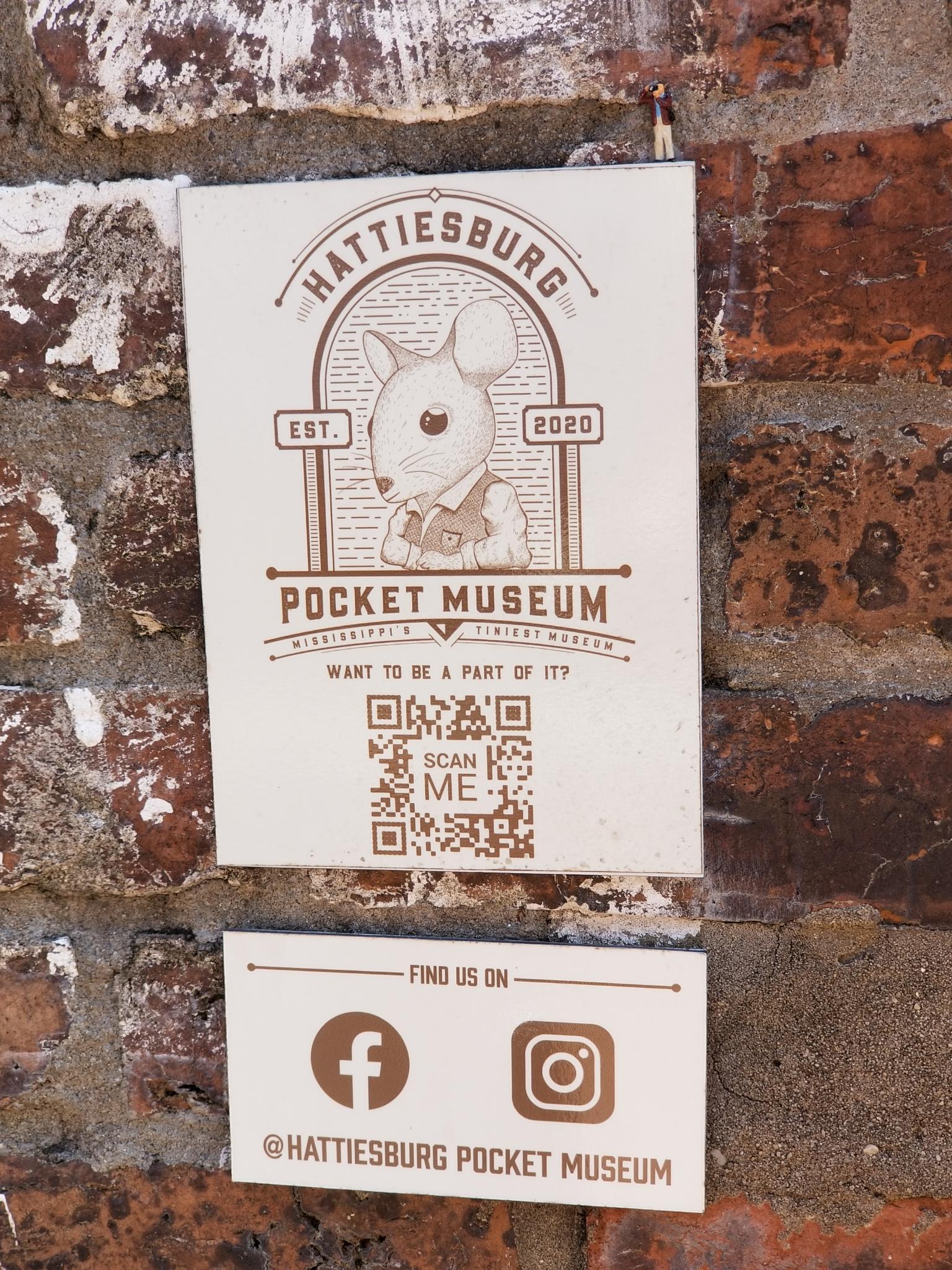 A sign on a brick wall marks the location of the 'Hattiesburg Pocket Museum, Mississippi's Tiniest Museum', and portrays a mouse wearing a vest.