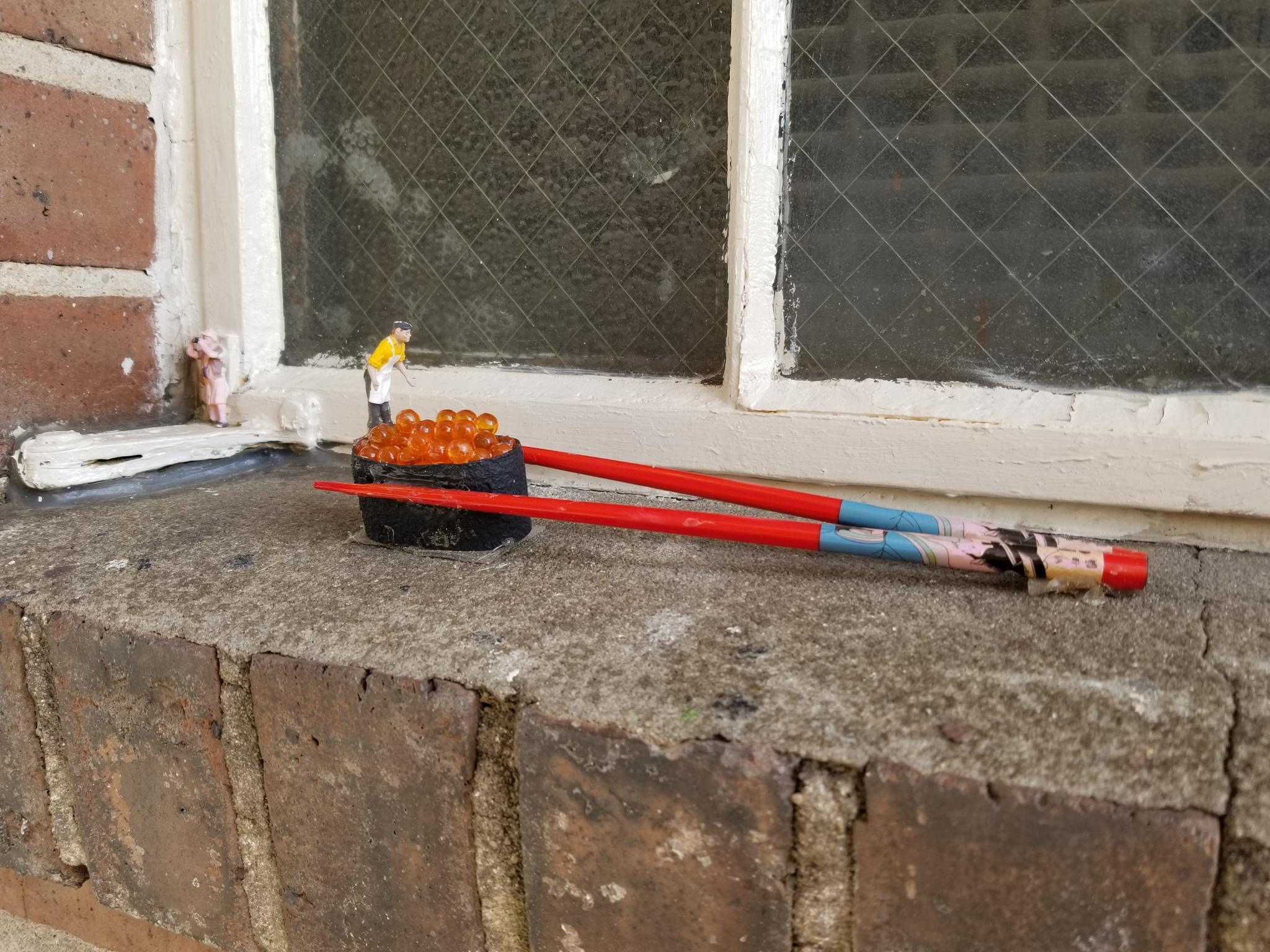 An art display on an exterior window sill in which a very small man appears to lean over a sushi roll held in place by chop sticks.