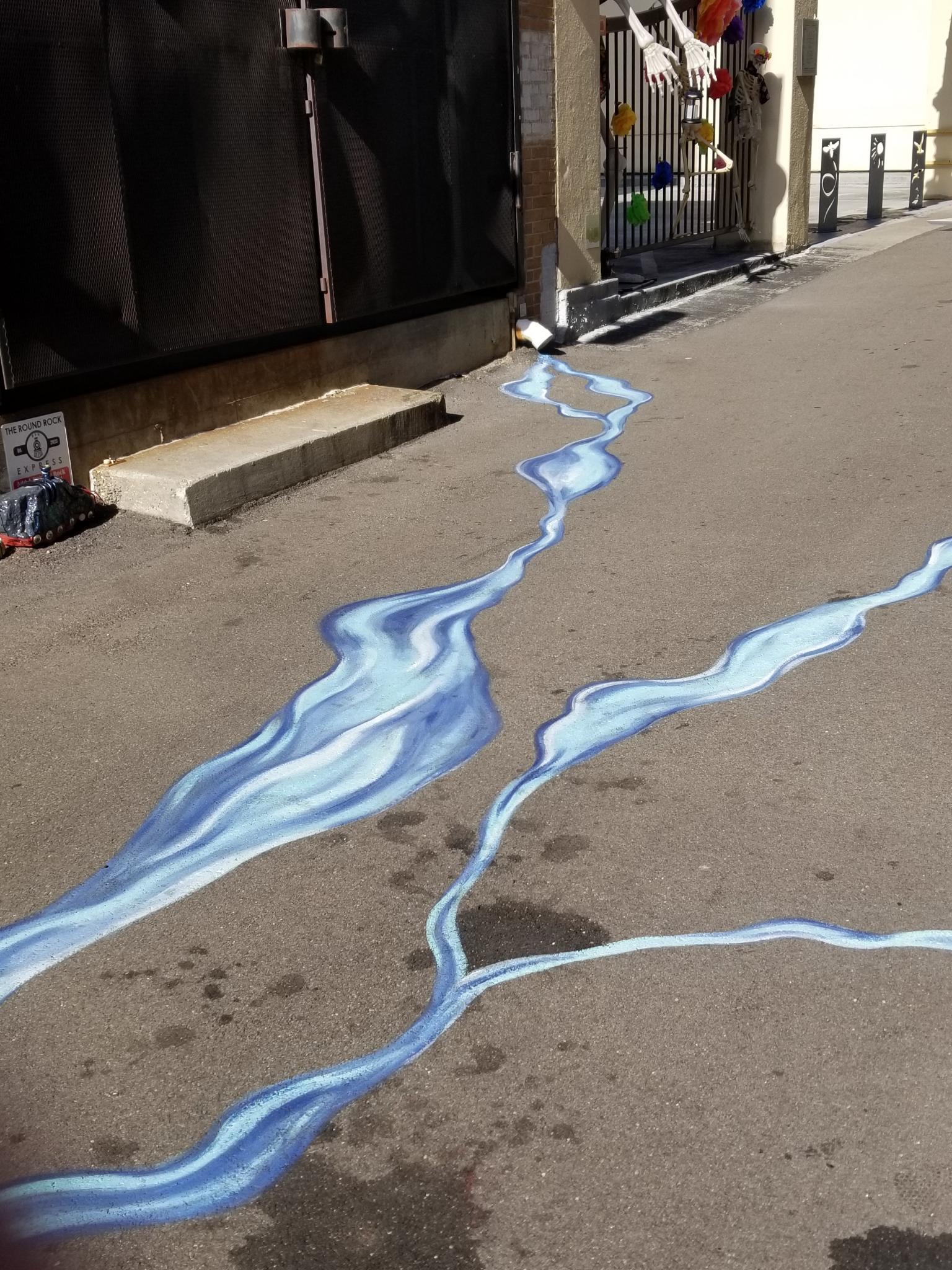 Painting on the ground of an alley that creates the appearance of water leaving a downspout and flowing along the alley.