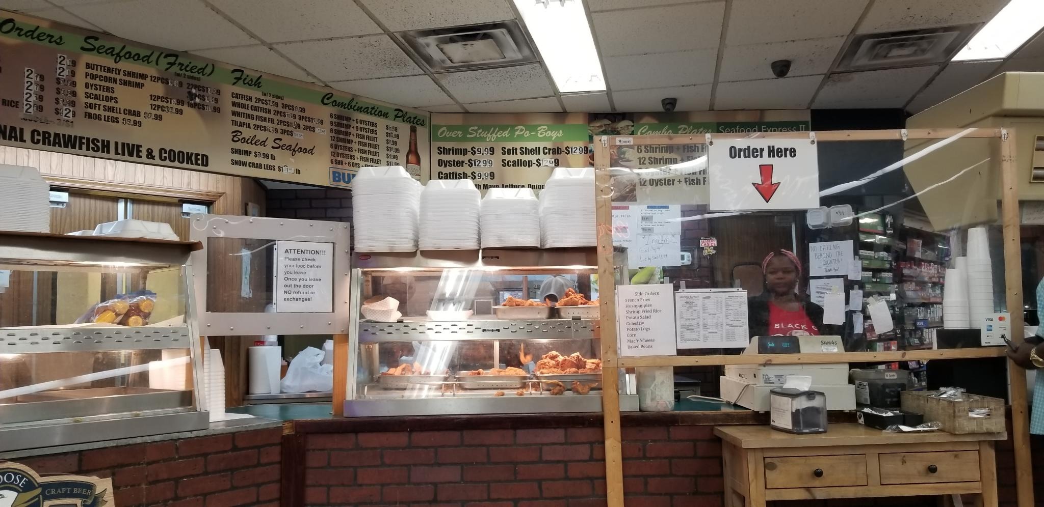 The order counter of a gas-station seafood restaurant where a woman awaits orders from under a sign that says 'Order Here'.