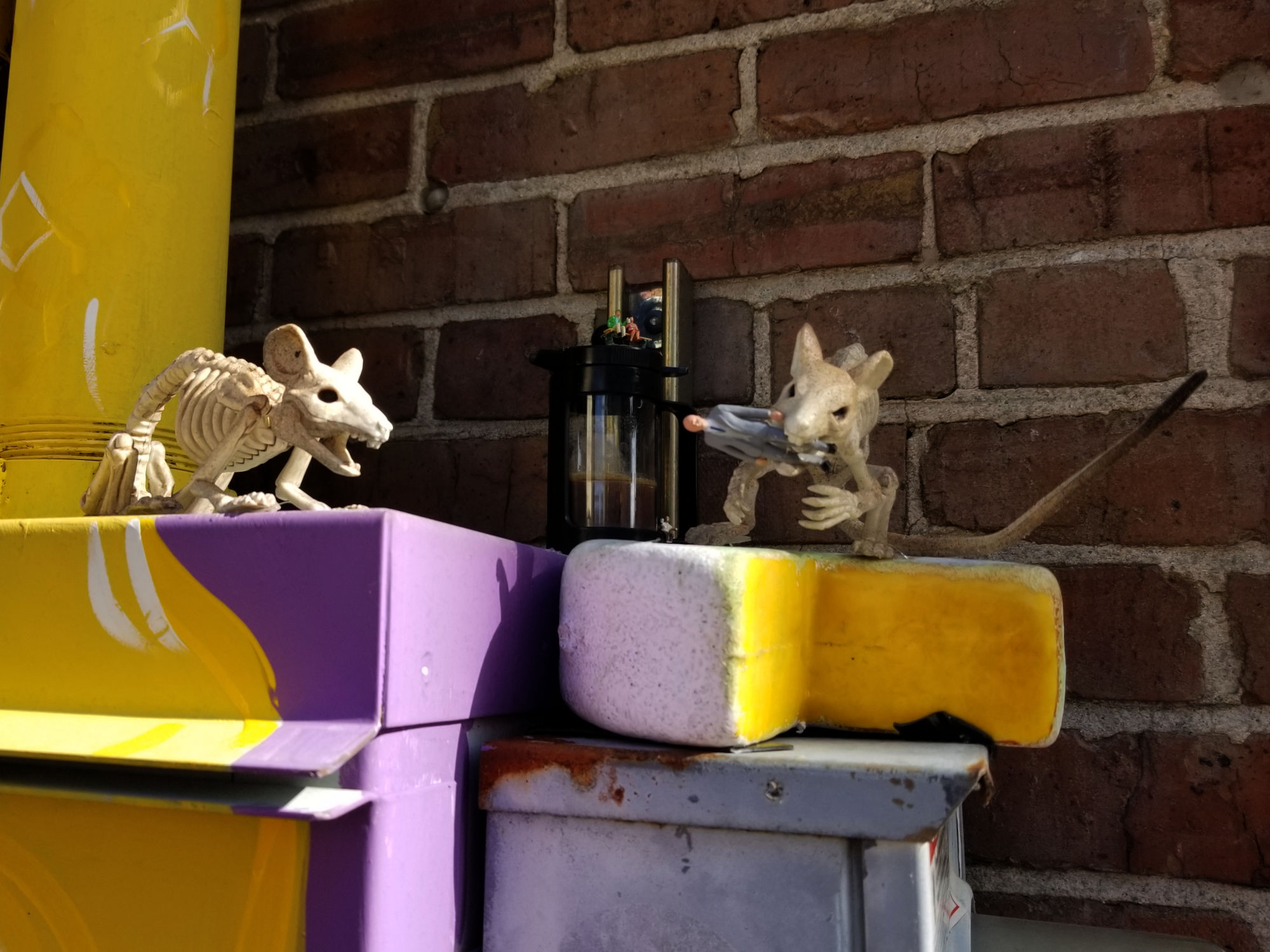 A small sculpture appears on top of some electrical boxes. A wheel of cheese missing a wedge. Two animated-seeming skeleton-mice, one of which hold a very small man in its jaws while two much smaller human figures look on.