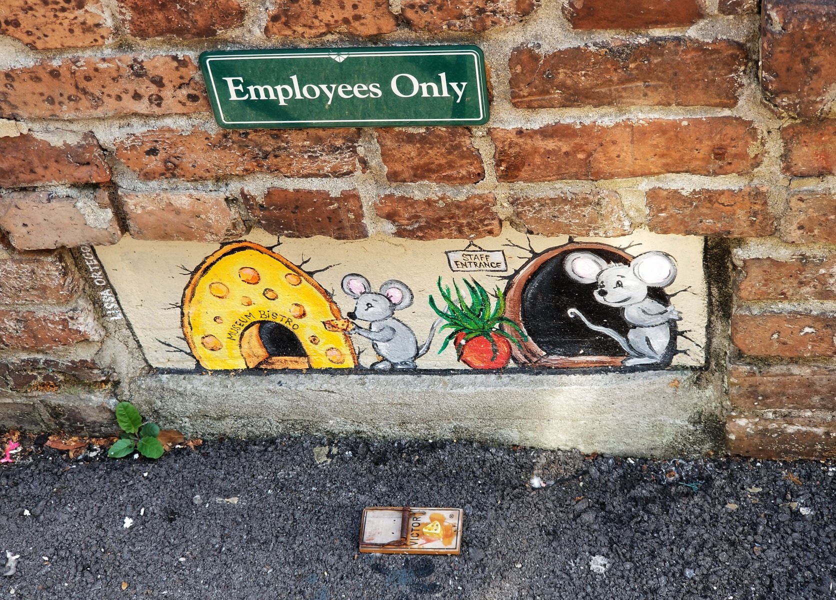 A painting in a section at the base of a brick wall showing two mice at the doors of what is indicated to be a bistro. On the ground outside is a mousetrap.