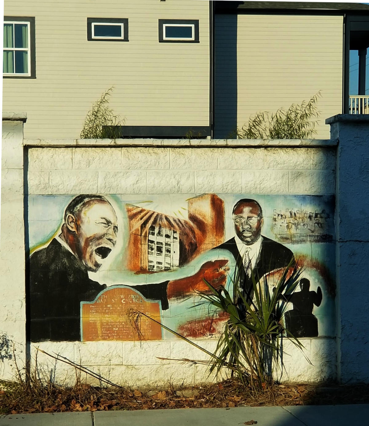 Mural on a white block wall portrays various images to include animated preaching.