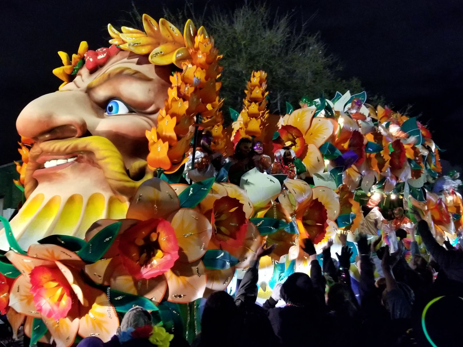 Parade float at night, covered with flowers and headed by a face with a prominent nose, golden laurel, and blonde mustache.