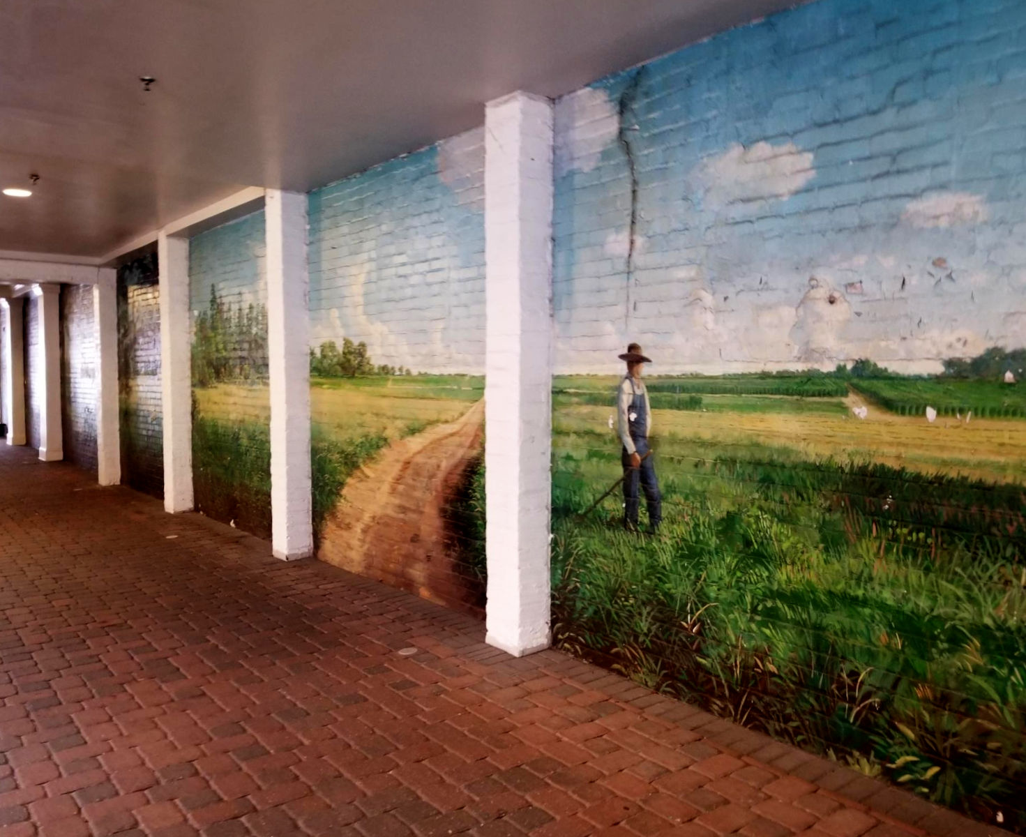 A painting spans the wall of a covered driveway, portraying a farmer standing his field into which a path appears to run from the driveway.