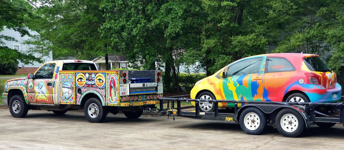 A car on a trailer bed behind a truck, but covered with art, which seems to have been professionally applied.