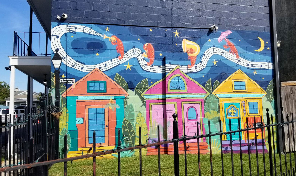 A mural covers the end of a residence portraying shrimp parading, playing music, along written music over top of colorful houses.