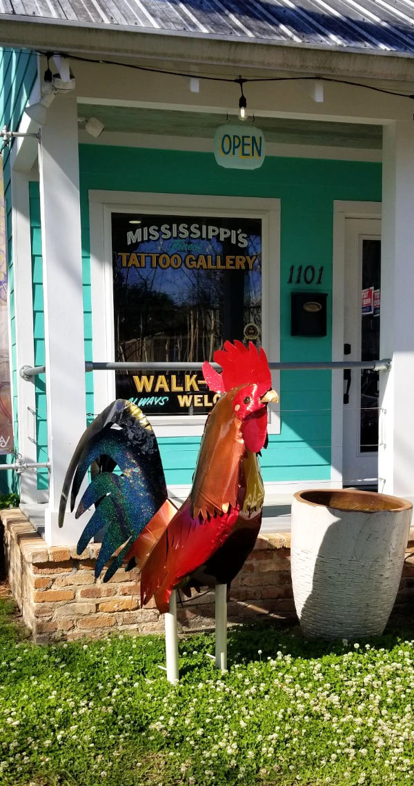 A tin sculpture made of time portraying a rooster, about three feet tall, standing on the grass in front of Mississippi's Tattoo Gallery (Walk-ins Always Welcome).