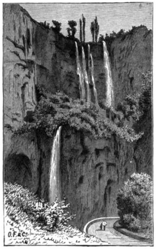 From a curving road, the viewer sees a waterfall comprising two sections.