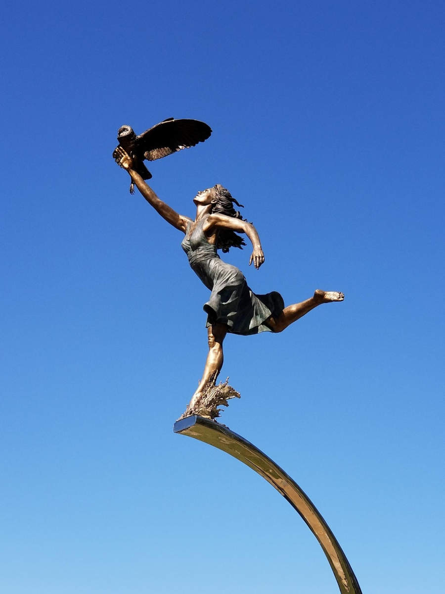 In front of a clear sky, a bronze statue, female form, right leg raised, left leg splashing, right arm raised, holding an owl that is taking flight.