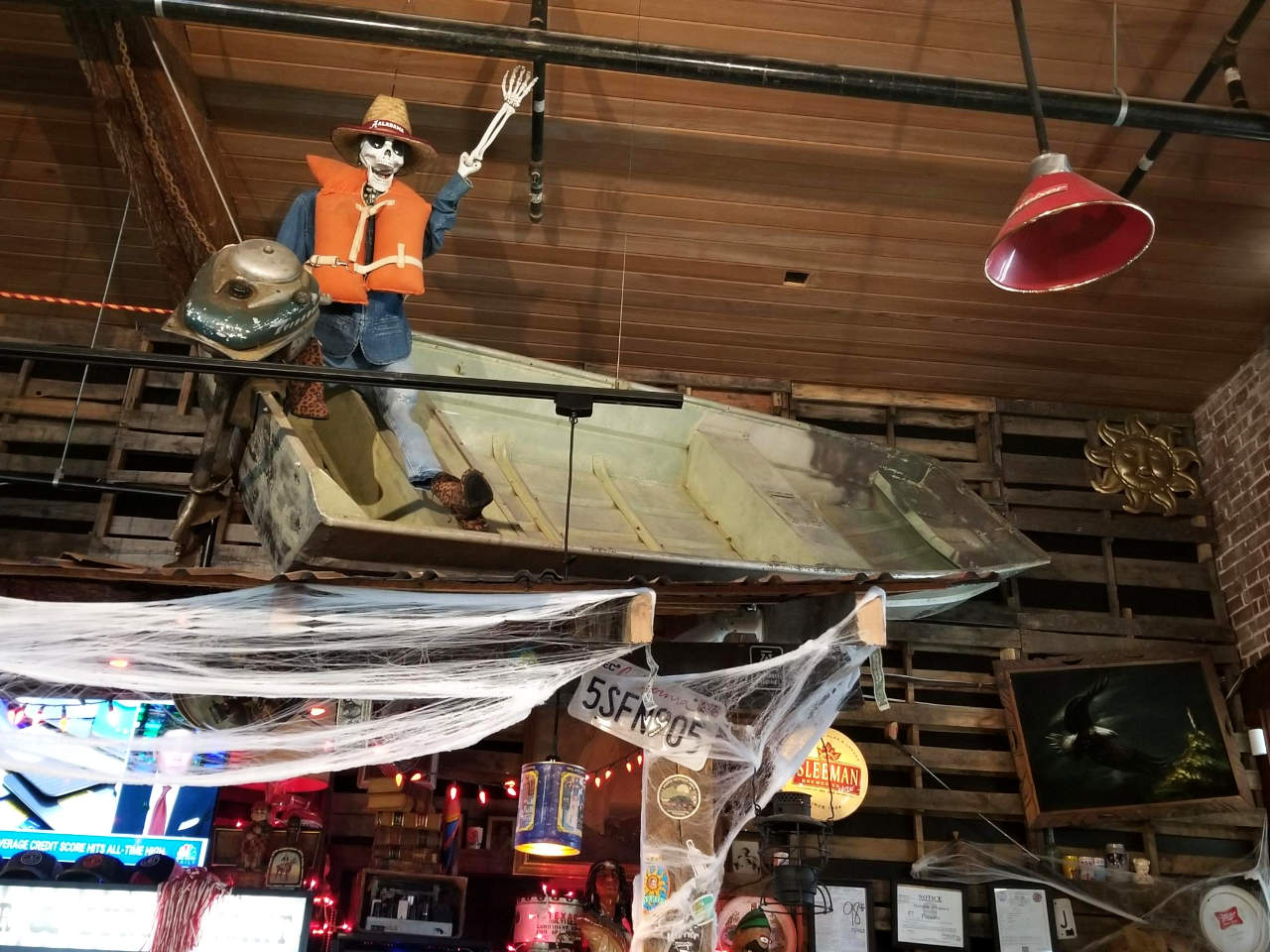 A small boat with an outboard motor, suspended from a restaurant ceiling, with a skeleton model seeming to steer, arm upraised, wearing clothing, a hat, sunglasses, and a life-jacket.