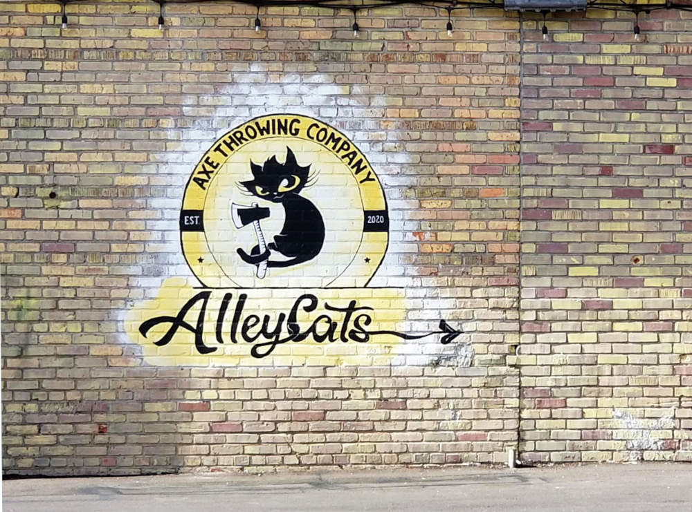 Murpentraĵo sur brika muro reklamanta la Alley Cats Axe Throwing Company kaj prezentanta nigran katidon kun grandaj okuloj tenante hakilon.