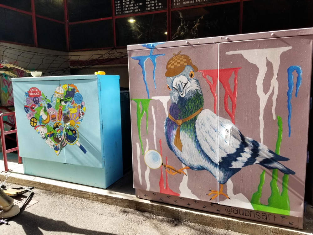 Paintings on metal boxes, a large pigeon wearing a hat and tie like Sherlock Holmes, holding a magnifying glass, with colorful splotches.