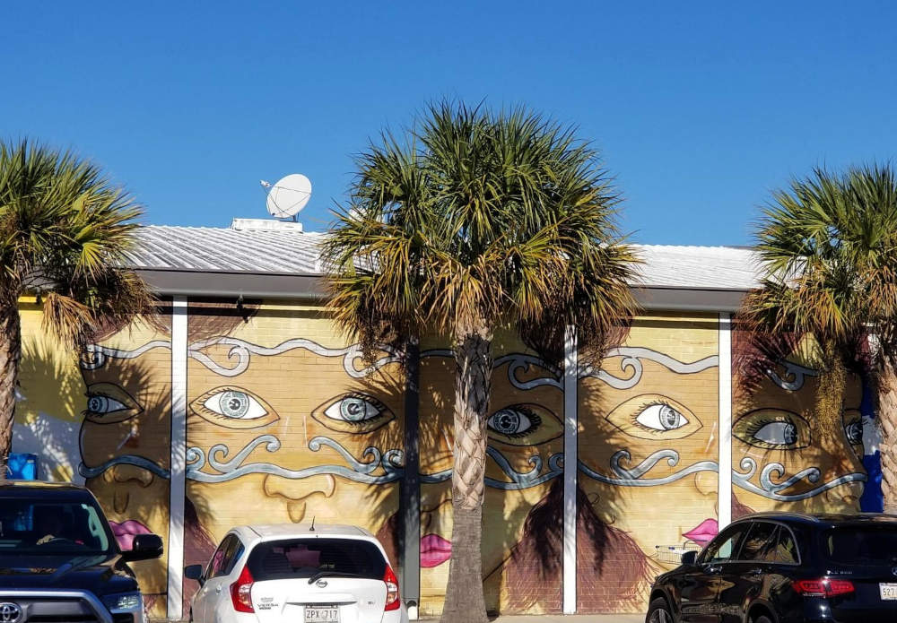 Malantaŭ palmoj, muro de konstrua kovrita per murpentraĵo montras vizaĝojn portantajn maskeradaj maskojn, kiuj ŝajnas gardi la parkejon.