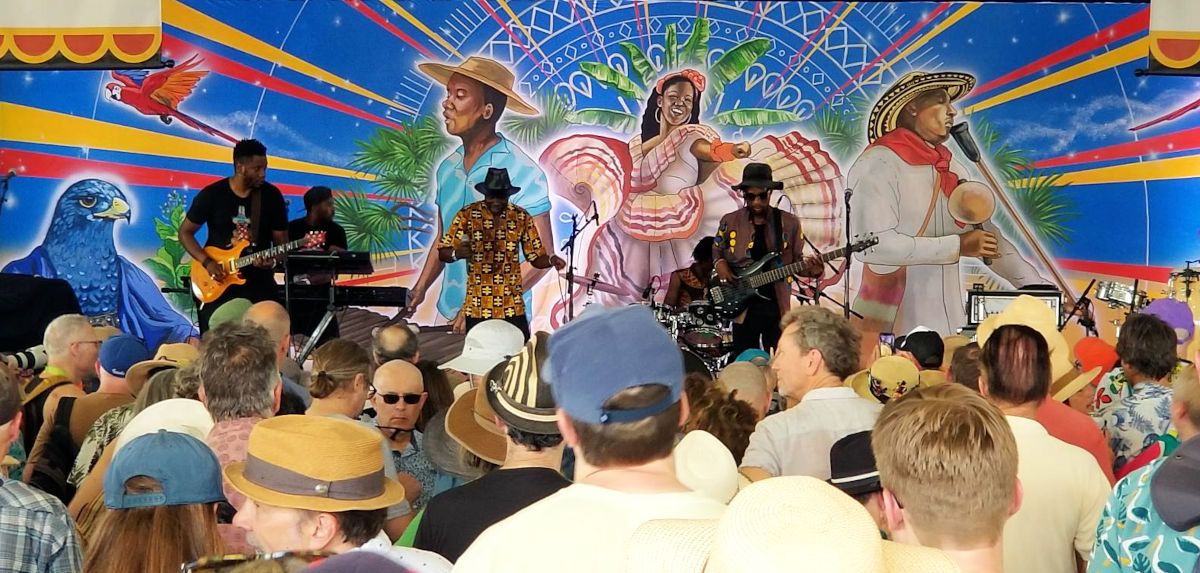 Kvar muzikistoj ludas sur scenejo, fronte al malgranda homamaso kaj antaŭ bunta afrika-tema bildo.