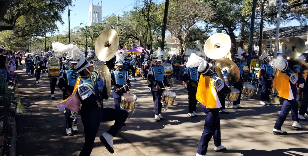 Three paraders playing cymbals, left cymbal upraised, drummers following.