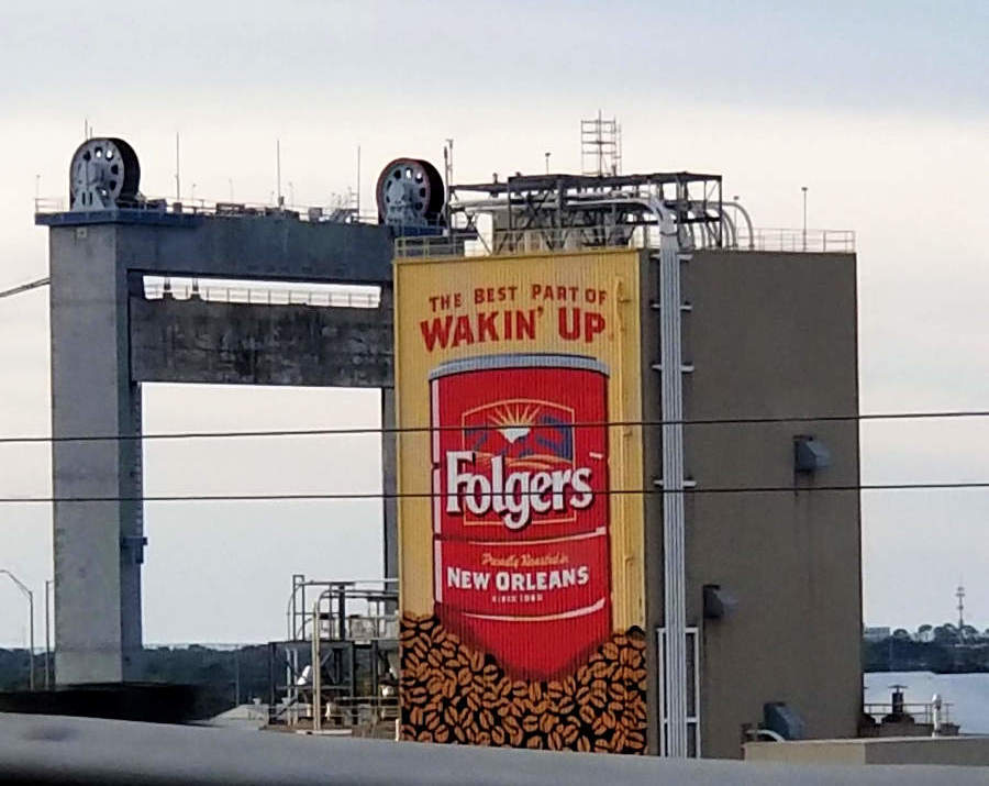 A large building in the distance, past a guard-rail, one side of which is covered by the image of a Folgers coffee can, an advertising slogan, and the message that the brand is made in New Orleans.