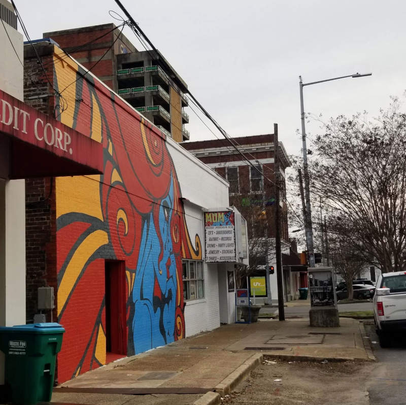 A mural next to a city street, a profile face of a blue-haired woman with red hair, crying black tears.