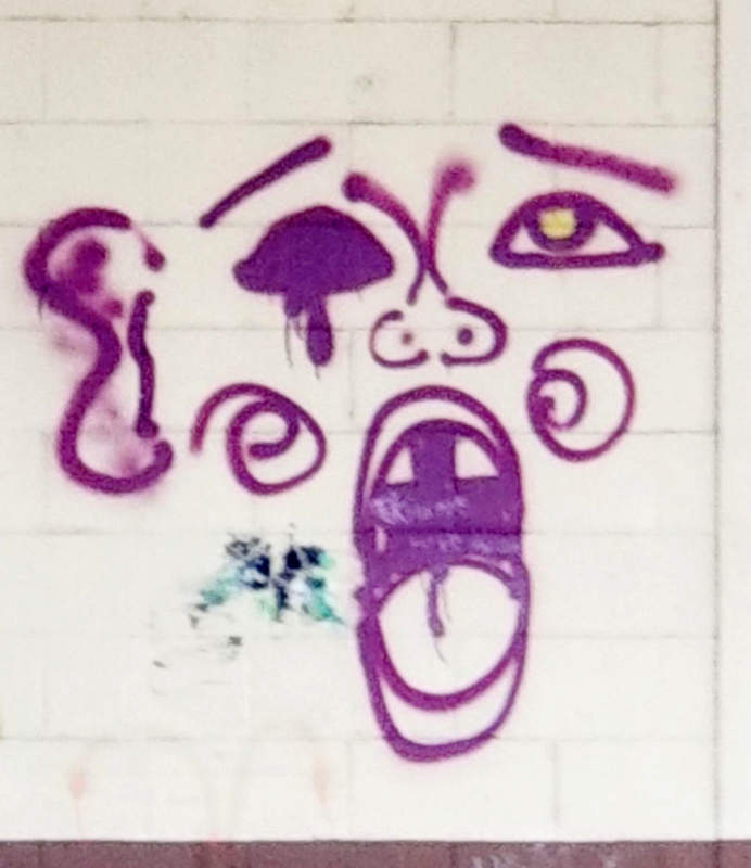 A rough image of a face, open mouth, one dark eye, drawn with purple spray paint on a wall.