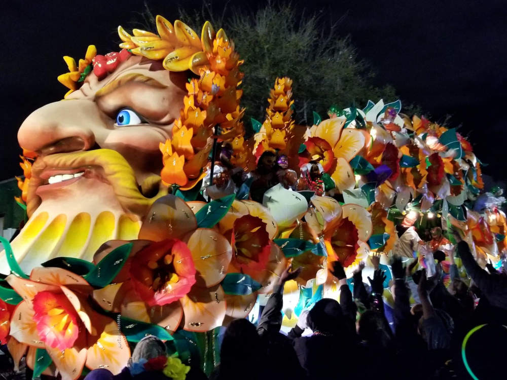 Parade float, at night, covered with flowers with a face, prominent nose, golden laurel, and blonde mustache.